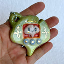 Ceramic sculpture of a green gingko leaf, modeled to be a keychain with buttons on the front like a tamagotchi toy. Inside the screen is a smaller tan gingko leaf with large eyes and a cute smile.
