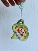 Ceramic sculpture of a green gingko leaf, modeled to be a keychain with buttons on the front like a tamagotchi toy. Inside the screen is a smaller tan gingko leaf with large eyes and a cute smile.