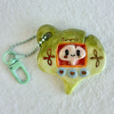 Ceramic sculpture of a green gingko leaf, modeled to be a keychain with buttons on the front like a tamagotchi toy. Inside the screen is a smaller tan gingko leaf with large eyes and a cute smile.