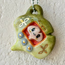 Ceramic sculpture of a green gingko leaf, modeled to be a keychain with buttons on the front like a tamagotchi toy. Inside the screen is a smaller tan gingko leaf with large eyes and a cute smile.