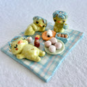 Set of several ceramic sculptures making up a picnic setting with 3 kappas dining outside on a plaid blanket with various Japanese food items and cups of green tea.