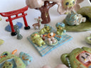 Set of several ceramic sculptures making up a picnic setting with 3 kappas dining outside on a plaid blanket with various Japanese food items and cups of green tea.
