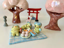 Set of several ceramic sculptures making up a picnic setting with 3 kappas dining outside on a plaid blanket with various Japanese food items and cups of green tea.