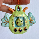 Glossy ceramic sculpture of a leaf themed Tamagotchi toy, with a small green leaf as the inner character and gold gilded patterning and buttons. On the side of the Tamamgotchi are leaf shaped wings and atop are orange antennae. 