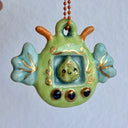 Glossy ceramic sculpture of a leaf themed Tamagotchi toy, with a small green leaf as the inner character and gold gilded patterning and buttons. On the side of the Tamamgotchi are leaf shaped wings and atop are orange antennae. 