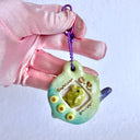 Glossy ceramic sculpture of a Tamagotchi toy with a green leaf shaped body and a small Kurchipatchi character inside. Gold gilded patterning and buttons are on the you and it is attached to a chain.