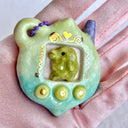 Glossy ceramic sculpture of a Tamagotchi toy with a green leaf shaped body and a small Kurchipatchi character inside. Gold gilded patterning and buttons are on the you and it is attached to a chain.