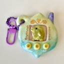 Glossy ceramic sculpture of a Tamagotchi toy with a green leaf shaped body and a small Kurchipatchi character inside. Gold gilded patterning and buttons are on the you and it is attached to a chain.