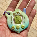 Glossy ceramic sculpture of a Tamagotchi toy with a green leaf shaped body and a small Kurchipatchi character inside. Gold gilded patterning and buttons are on the you and it is attached to a chain.