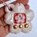 Glossy ceramic sculpture of a Tamagotchi toy with a pink cherry blossom shaped body and a small cherry blossom character inside. Gold gilded patterning and buttons are on the toy and it is attached to a chain.