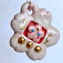 Glossy ceramic sculpture of a Tamagotchi toy with a pink cherry blossom shaped body and a small cherry blossom character inside. Gold gilded patterning and buttons are on the toy and it is attached to a chain.