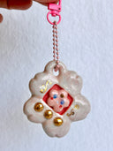 Glossy ceramic sculpture of a Tamagotchi toy with a pink cherry blossom shaped body and a small cherry blossom character inside. Gold gilded patterning and buttons are on the toy and it is attached to a chain.