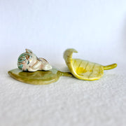 Glossy ceramic sculpture of a small fairy with short blue and gold hair, laying inside of a bed made of 2 leaves, with the top leaf curled over slightly like a blanket.