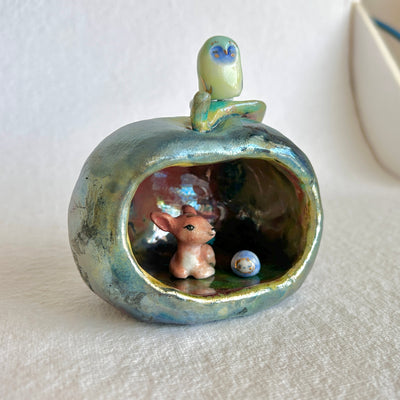 Ceramic sculpture of an apple, hollowed out on one side to make a cave of sorts. A small deer resides in it with a smaller round blue and white blob character with a sweet face.