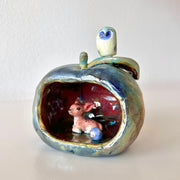 Ceramic sculpture of an apple, hollowed out on one side to make a cave of sorts. A small deer resides in it with a smaller round blue and white blob character with a sweet face.
