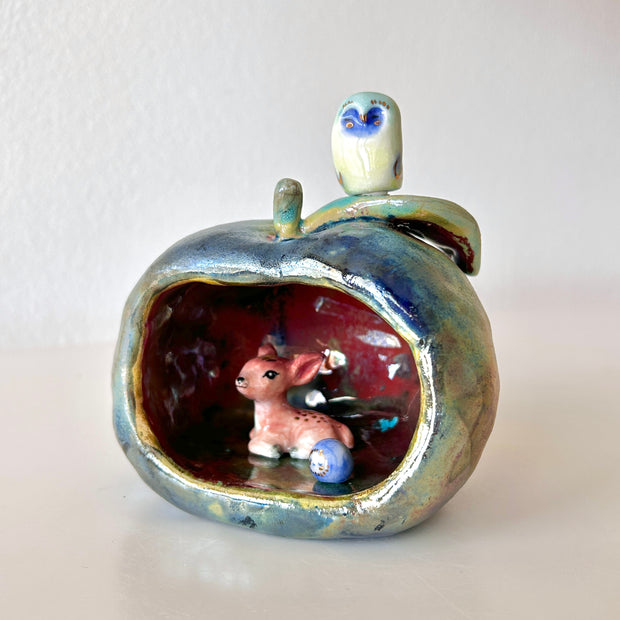 Ceramic sculpture of an apple, hollowed out on one side to make a cave of sorts. A small deer resides in it with a smaller round blue and white blob character with a sweet face.