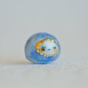 Small character ceramic sculpture of a orb with blue body and a tiny white face with gold cheeks and gold dots surrounding its face.