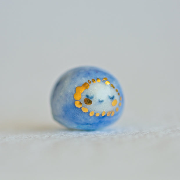 Small character ceramic sculpture of a orb with blue body and a tiny white face with gold cheeks and gold dots surrounding its face.