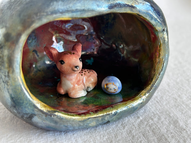 Ceramic sculpture of an apple, hollowed out on one side to make a cave of sorts. A small deer resides in it with a smaller round blue and white blob character with a sweet face.