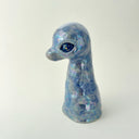 A small sculptural figurine shaped like a diplosaurus neck and head, covered in an iridescent blue, purple, and green glaze with gold foil flecks, photographed against a plain light background.