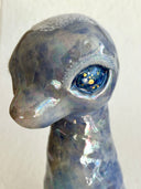 A small sculptural figurine shaped like a diplosaurus neck and head, covered in an iridescent blue, purple, and green glaze with gold foil flecks, photographed against a plain light background.