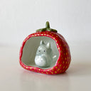 Small ceramic sculpture of a wide strawberry, hollowed out on one side as if it is a cave of sorts. A small sculpture of Totoro sits inside.