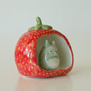Small ceramic sculpture of a wide strawberry, hollowed out on one side as if it is a cave of sorts. A small sculpture of Totoro sits inside.
