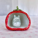 Small ceramic sculpture of a wide strawberry, hollowed out on one side as if it is a cave of sorts. A small sculpture of Totoro sits inside.
