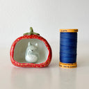 Small ceramic sculpture of a wide strawberry, hollowed out on one side as if it is a cave of sorts. A small sculpture of Totoro sits inside. Piece sits next to a spool of thread for size reference.