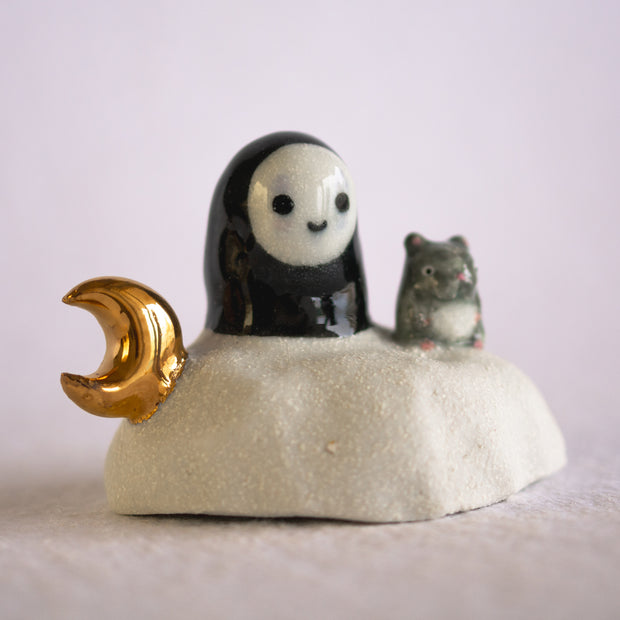 Ceramic sculpture of a No Face character sitting on a rock-like mound with a tiny mouse next to it and a golden moon on its other side.