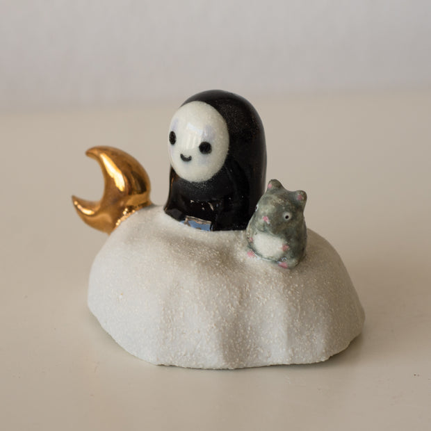 Ceramic sculpture of a No Face character sitting on a rock-like mound with a tiny mouse next to it and a golden moon on its other side.