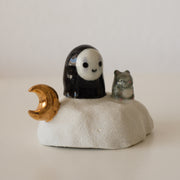 Ceramic sculpture of a No Face character sitting on a rock-like mound with a tiny mouse next to it and a golden moon on its other side.