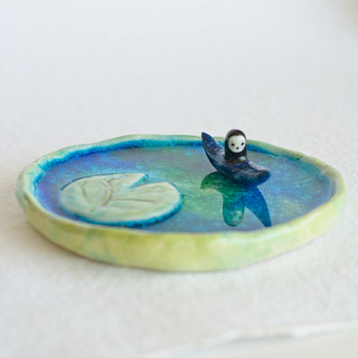 Ceramic sculpture of a lotus pond, with a single large lotus leaf on the water's surface and a small character in a kayak.