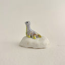 A small decorative figurine of a loch ness monster, sitting on a white rock, with a pale lavender-gray body, yellow accents, and gold glitter speckles, set against a plain light beige background.