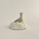 A small decorative figurine of a loch ness monster, sitting on a white rock, with a pale lavender-gray body, yellow accents, and gold glitter speckles, set against a plain light beige background.