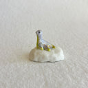 A small decorative figurine of a loch ness monster, sitting on a white rock, with a pale lavender-gray body, yellow accents, and gold glitter speckles, set against a plain light beige background.