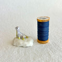 A small decorative figurine of a loch ness monster, sitting on a white rock, with a pale lavender-gray body, yellow accents, and gold glitter speckles, set against a plain light beige background.