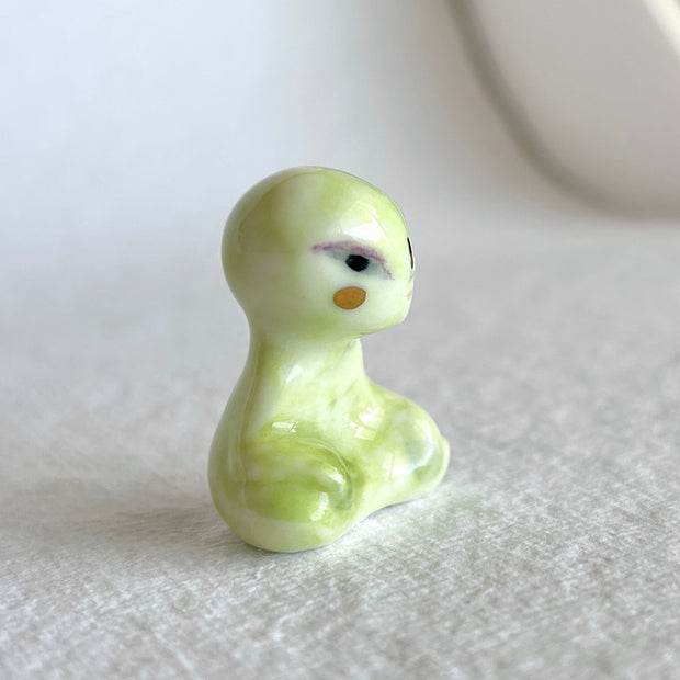 Small ceramic sculpture of a green alien, sitting in a meditation pose with crossed legs.