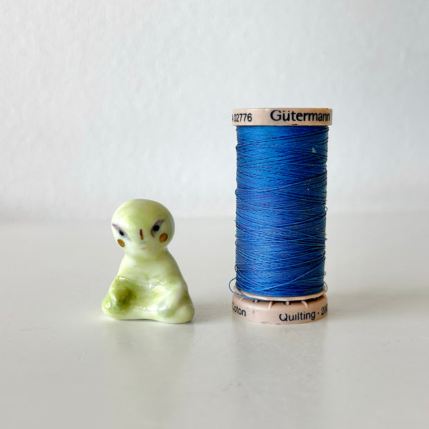 Small ceramic sculpture of a green alien, sitting in a meditation pose with crossed legs. Next to a spool of thread for size reference.