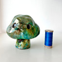 Raku fired ceramic sculpture of a piece of broccoli, with a cloud shaped top, a set of black eyes and a tiny stem for a body. Next to a spool of thread for size reference.