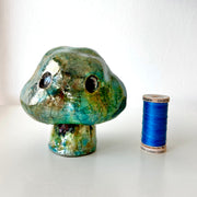 Raku fired ceramic sculpture of a piece of broccoli, with a cloud shaped top, a set of black eyes and a tiny stem for a body. Next to a spool of thread for size reference.