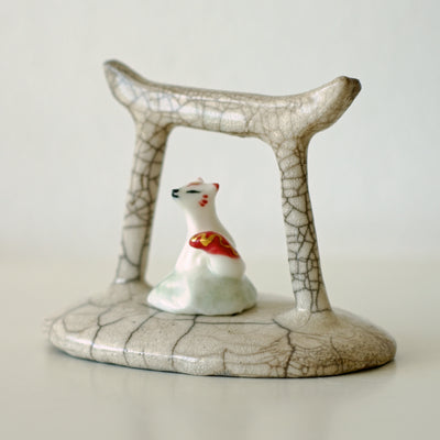 Ceramic sculpture of a Torii gate in a muted beige color with a white fox spirit sitting in between them. He has red tipped ears and a red and gold sash around him.