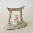 Ceramic sculpture of a Torii gate in a muted beige color with a white fox spirit sitting in between them. He has red tipped ears and a red and gold sash around him.