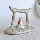 Ceramic sculpture of a Torii gate in a muted beige color with a white fox spirit sitting in between them. He has red tipped ears and a red and gold sash around him.