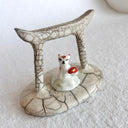 Ceramic sculpture of a Torii gate in a muted beige color with a white fox spirit sitting in between them. He has red tipped ears and a red and gold sash around him.