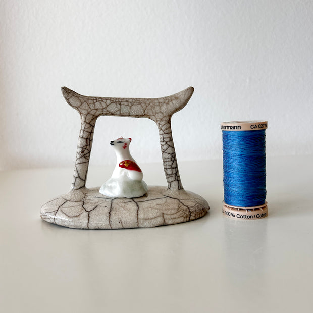 Ceramic sculpture of a Torii gate in a muted beige color with a white fox spirit sitting in between them. He has red tipped ears and a red and gold sash around him.