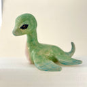 A small stylized sea creature figurine, resembling a type of baby water dinosaur, made from translucent green and blue ceramics with a glittery black eye, resting on a white surface against a light beige background.