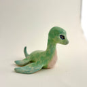 A small stylized sea creature figurine, resembling a type of baby water dinosaur, made from translucent green and blue ceramics with a glittery black eye, resting on a white surface against a light beige background.