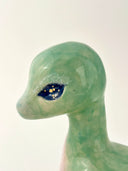 A small stylized sea creature figurine, resembling a type of baby water dinosaur, made from translucent green and blue ceramics with a glittery black eye, resting on a white surface against a light beige background.