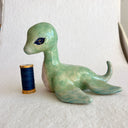 A small stylized sea creature figurine, resembling a type of baby water dinosaur, made from translucent green and blue ceramics with a glittery black eye, resting on a white surface against a light beige background.